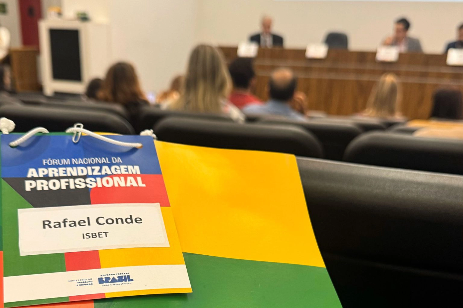 na foto: pasta com o tema do fórum de aprendizagem profissional em foco, na frente a reunião.