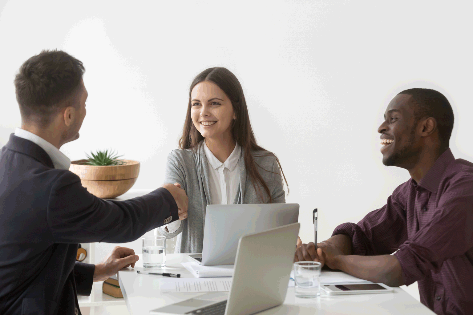 Na mesa tem três jovens sentados em uma entrevista de emprego para jovem aprendiz, a garota foi aceita e está sorrindo apertando a mão de um dos rapazes o outro está feliz e sorrindo muito também.