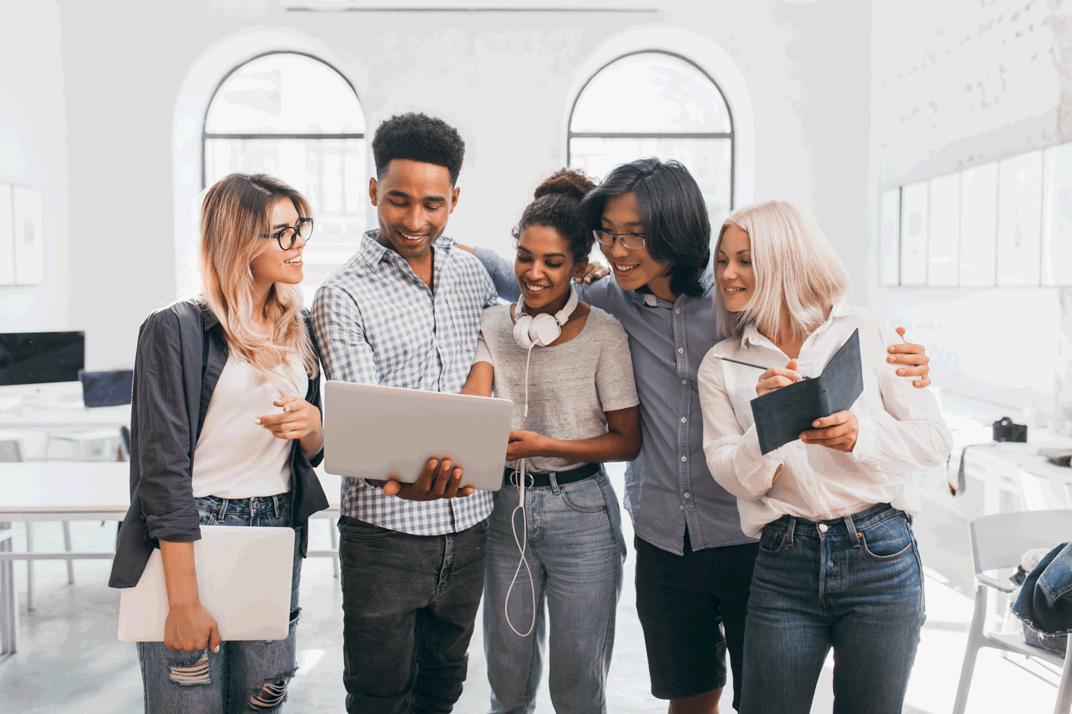 5 jovens de pé, olhando para um jovem que está segurando um notebook aberto. Alguns fazem anotações. Todos estão dentro do programa de estágio.