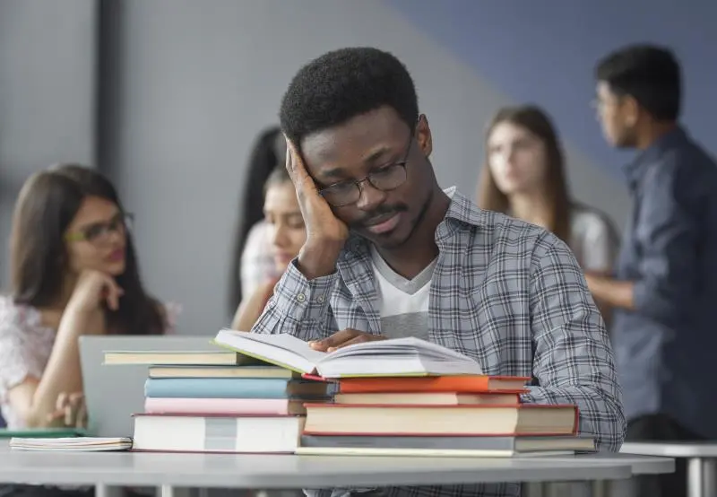 Jovem vestindo uma camisa xadrez, usando óculos, descansa a cabeça na palma da mão enquanto folheia um livro.