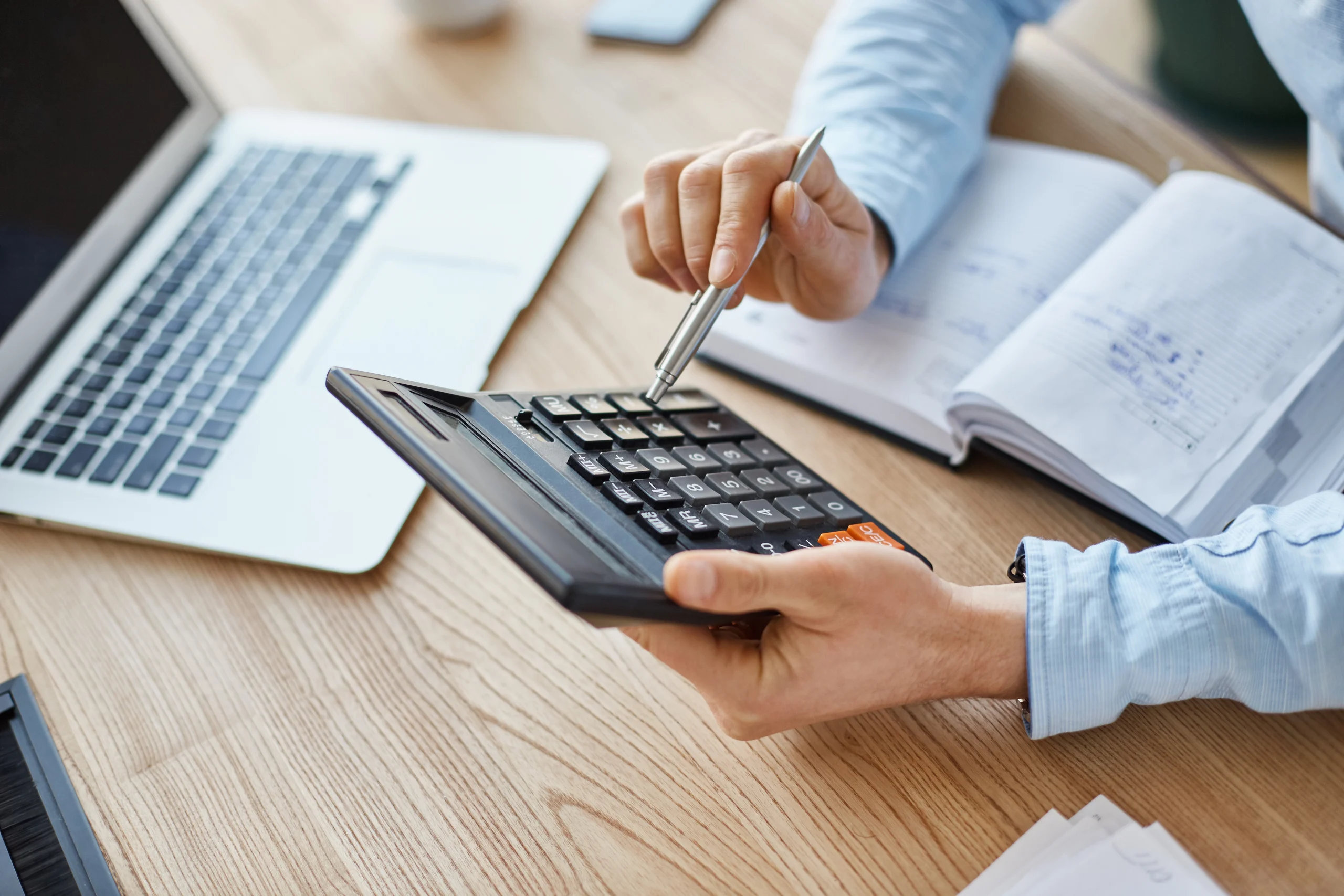 Na imagem uma mão segura uma calculadora, enquanto a outra usa uma caneta para selecionar a operação na calculadora, ambos os braços estão apoiados em uma mesa, com uma agenda e notebook abertos. A ideia é fazer alusão ao cálculo do salário do jovem aprendiz.