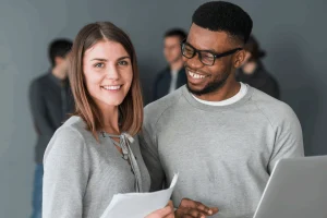 na imagem: dois estudantes estagiários sorriem um para o outro, um rapaz negro de óculos e uma jovem branca de cabelo chanel.