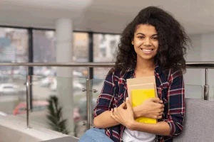 Na imagem uma jovem aprendiz de pé, segurando um caderno amarelo e envelopes, é uma jovem negra, com um bonito cabelo cacheado.