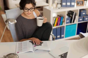 Na imagem uma jovem com uma caneca de café na mão, lê alguns livros e estuda um pouco!