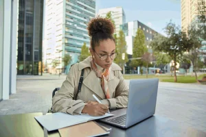 Na imagem uma estudante jovem, de pele negra, copia a matéria estudada do notebook para o caderno.