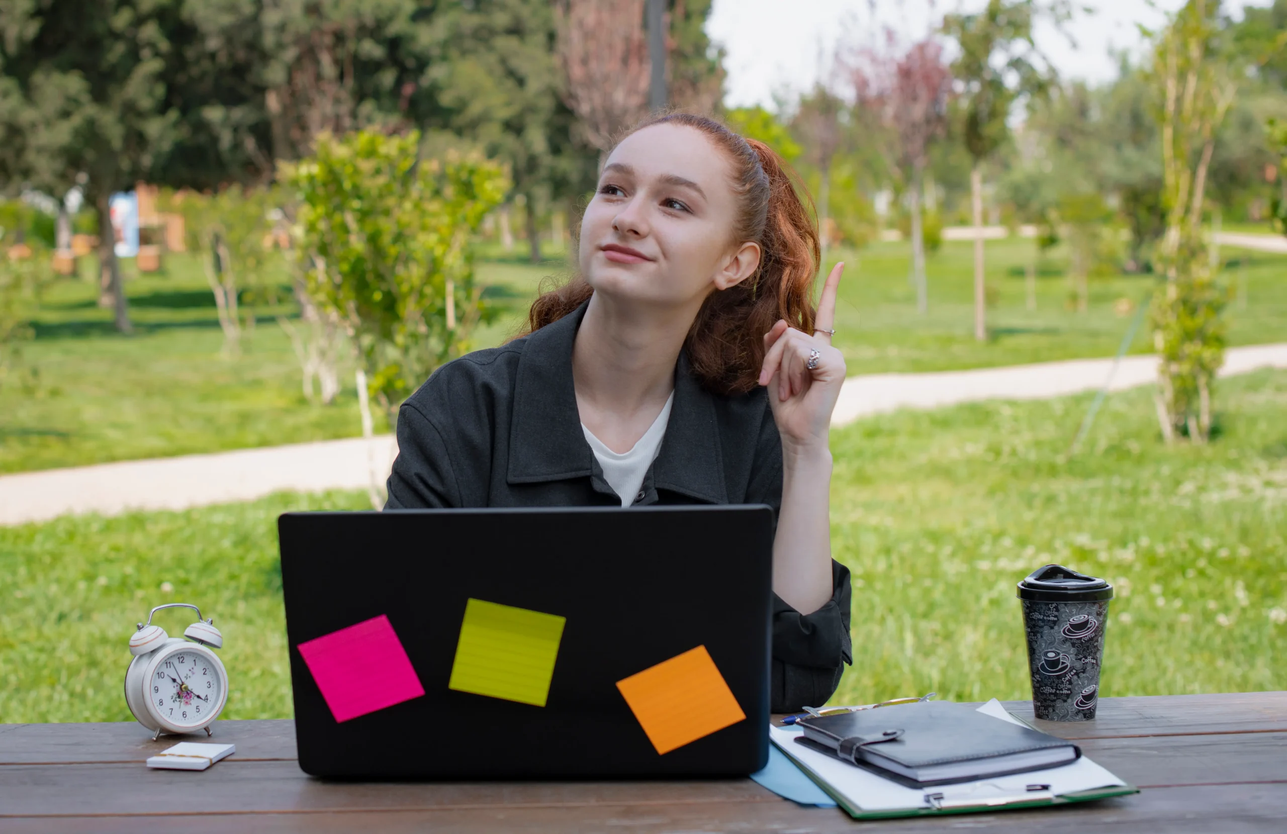Nesta imagem, uma jovem estudante de cabelos ruivos está sentada em uma mesa ao ar livre em um parque, usando um laptop com post-its coloridos colados na tampa. Ela parece pensativa, com o dedo levantado como se tivesse acabado de ter uma ideia. Ao redor dela, há materiais de estudo, um despertador branco e um copo de café, compondo um cenário de concentração criativa em meio à natureza. Essa imagem é perfeita para ilustrar temas como estudos ao ar livre, ensino remoto, criatividade, foco nos estudos e bem-estar acadêmico.