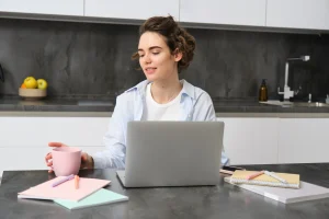 A imagem mostra uma mulher jovem sentada em uma cozinha moderna, utilizando um laptop. À sua frente, há cadernos coloridos, canetas e uma caneca rosa, indicando um ambiente de estudo organizado e acolhedor. Ela parece estar concentrada em um curso online ou atividade de trabalho remoto, transmitindo temas como educação digital, rotina de estudos, home office e autodisciplina. Ideal para ilustrar conteúdos sobre ensino a distância, organização pessoal, produtividade ou bem-estar no trabalho/studo em casa.