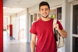 A imagem retrata um jovem universitário sorridente, vestindo uma camiseta vermelha e carregando uma mochila clara, enquanto está parado em um corredor escolar moderno. A luz natural e o ambiente limpo contribuem para uma atmosfera acolhedora e motivadora. Essa cena é ideal para ilustrar temas como educação superior, retorno às aulas, vida universitária, jovens no ensino médio ou técnico, e campanhas institucionais de universidades ou escolas.