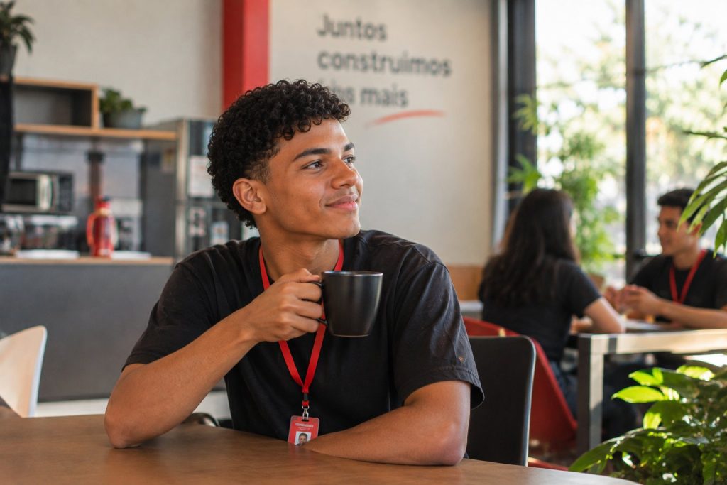 Jovem aprendiz com crachá vermelho fazendo uma pausa tranquila com uma xícara de café na área de descanso da empresa