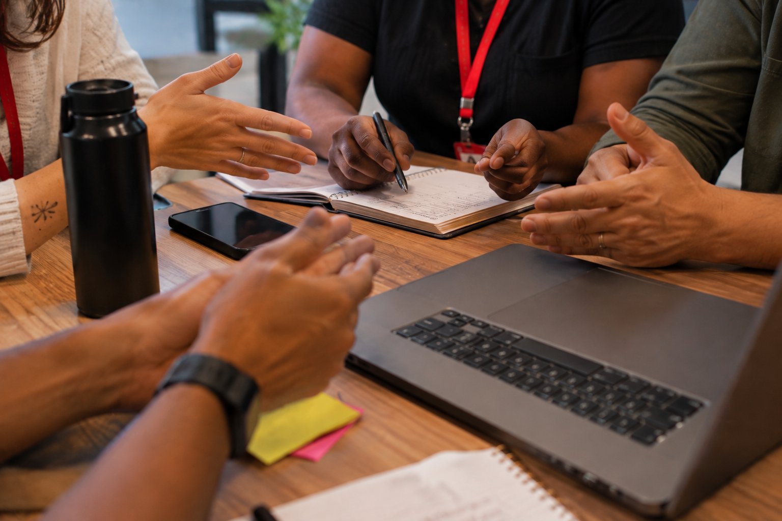 Equipe diversa reunida à mesa de trabalho discutindo estratégia, mãos gesticulando sobre anotações e notebook