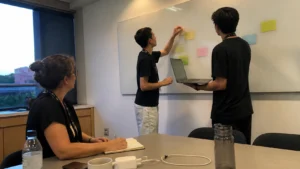 Jovens aprendizes montando um quadro de ideias com post-its enquanto uma colega acompanha, ilustrando o futuro do trabalho