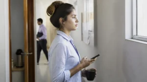 Jovem profissional faz uma pausa na janela do corredor para demonstrar maturidade emocional no trabalho antes de responder uma mensagem