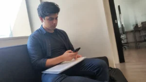 Jovem sentado no sofá com uma pasta no colo, conferindo no celular os requisitos para ser jovem aprendiz