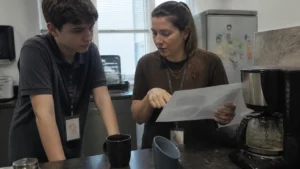 Soft skills no dia a dia, jovem aprendiz ouve atento a colega que explica algo em uma folha impressa na copa do escritório
