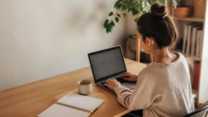 Jovem profissional trabalhando em notebook em um cantinho de home office aconchegante, com xícara de café e caderno de anotações